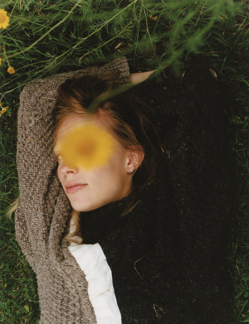 a woman laying in the grass wears a knit sweater, looking to camera as a flower obscures her eye