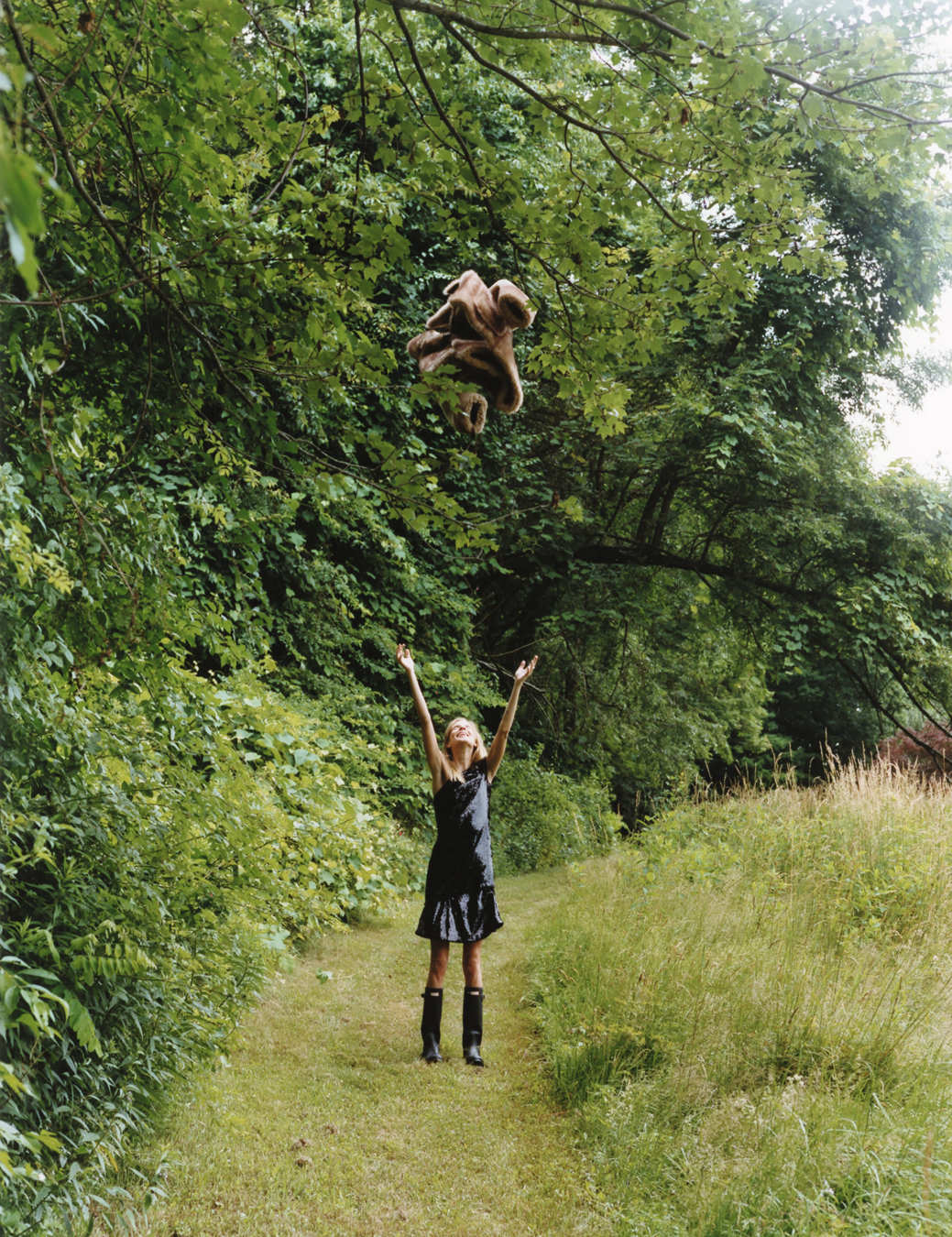 a woman in a black sequinned dress and wellies stands in the countryside, throwing her brown fur coat into the air above her head