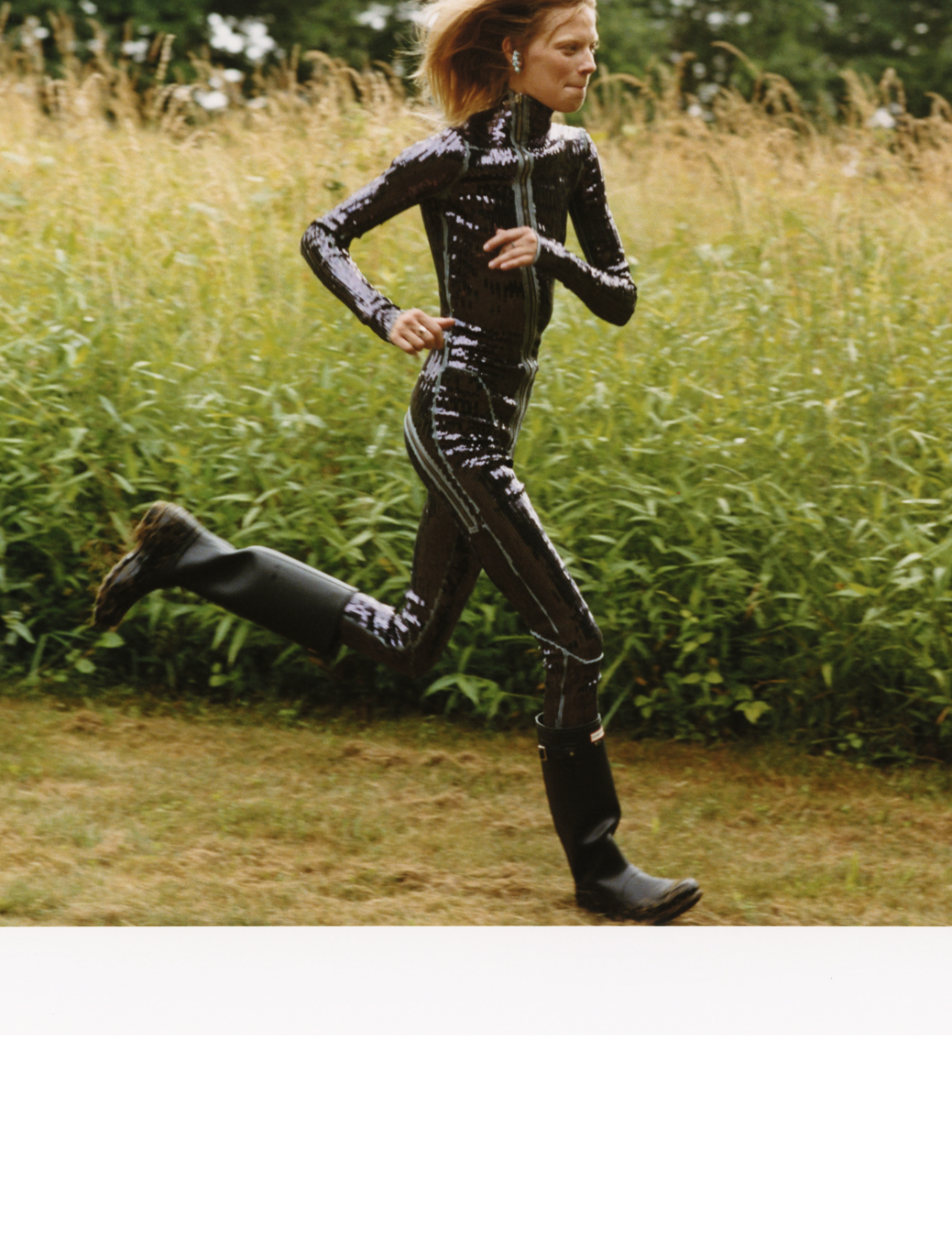 a woman in a black sequinned bodysuit and wellies runs through the countryside