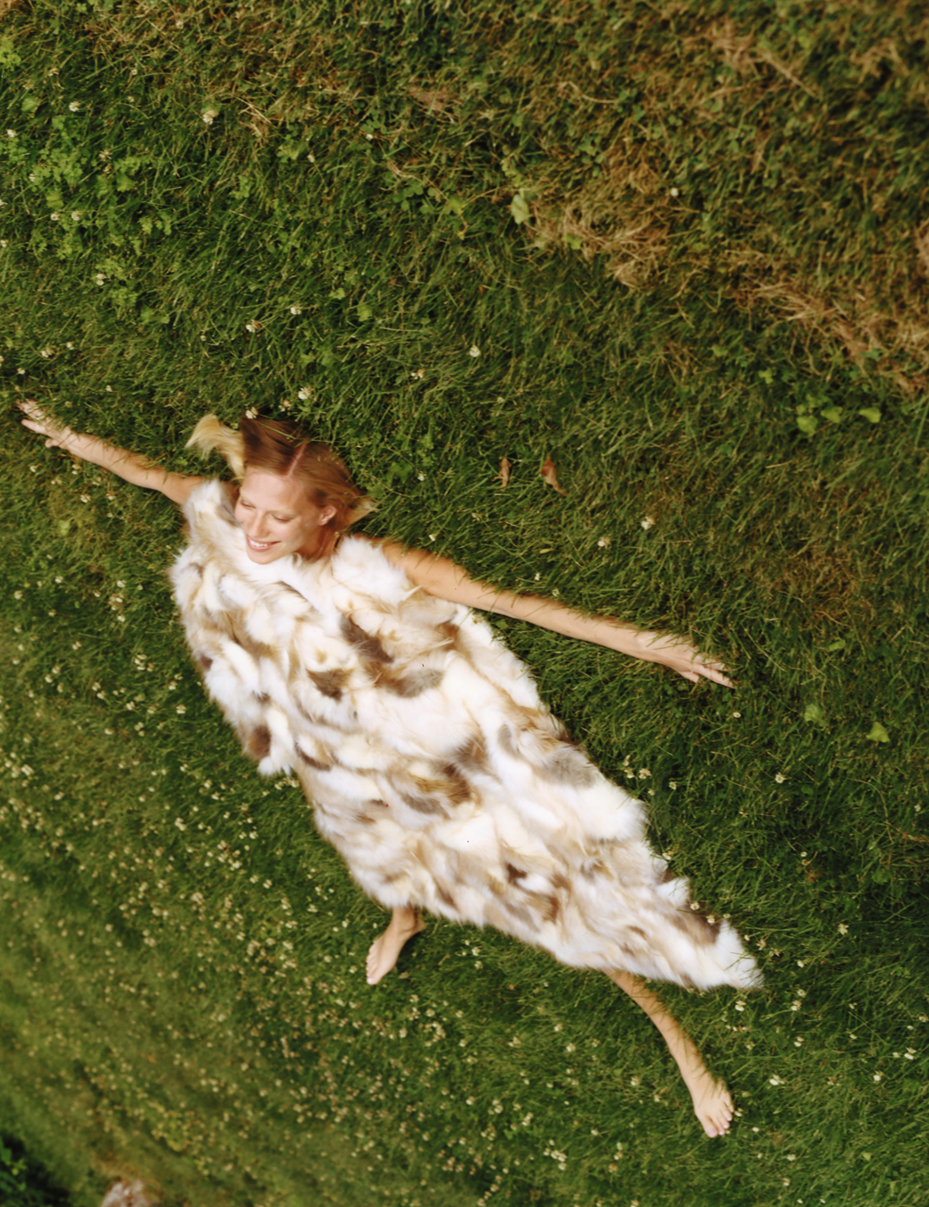 a woman lays in the grass with arms outstretched, a fur coat resting over her body