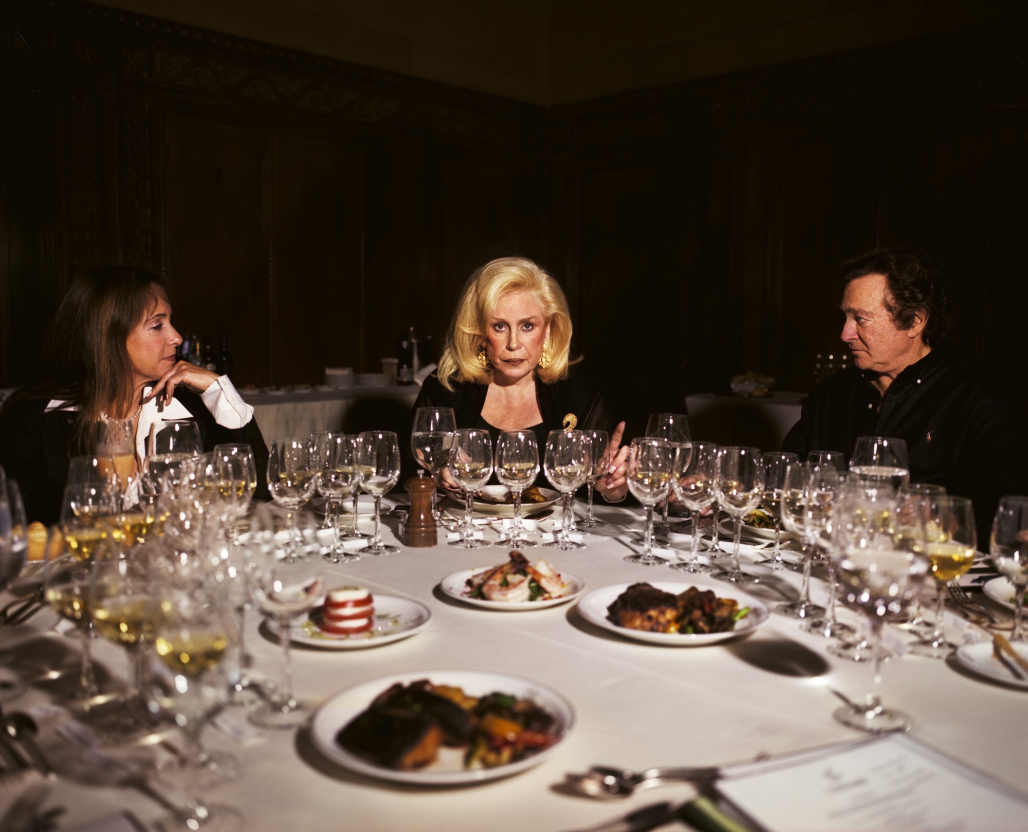 a blonde woman sits in the centre surrounded by glasses and plates, with a man and a woman to each side