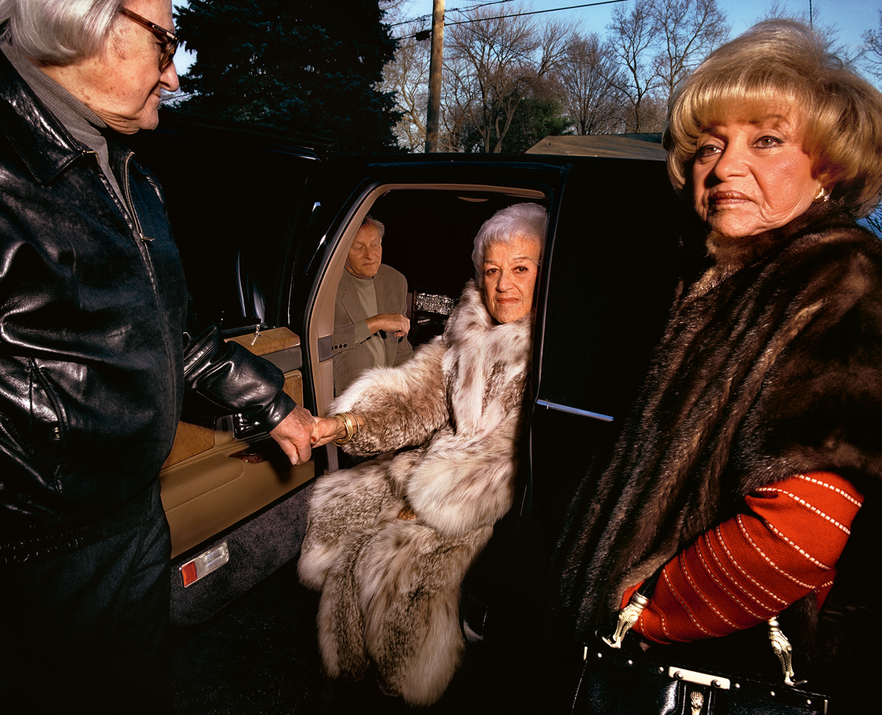 A woman in a fure coat is helped out the backseat of a car