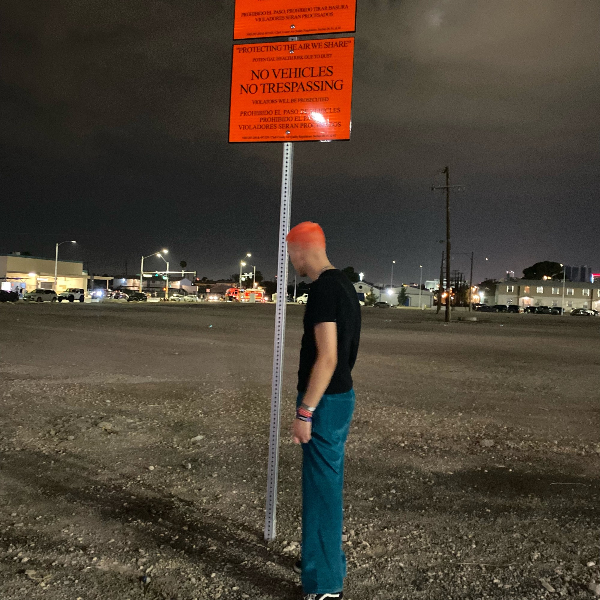 A man stands in front of a sign saying no vehicles no trespassing in the middle of a barren space at night.