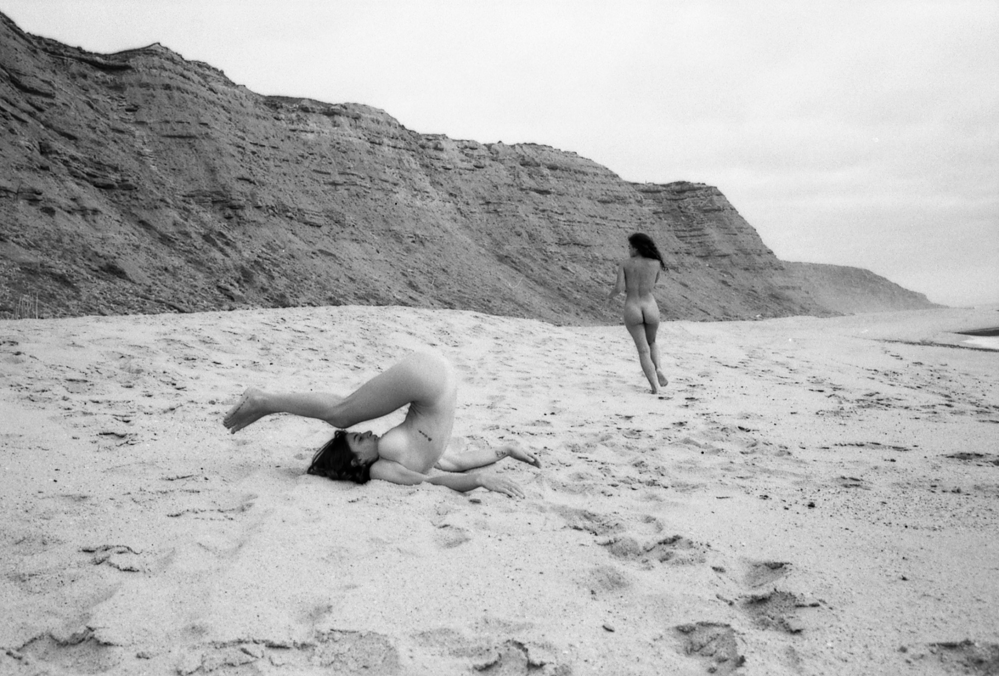 Two naked women play and run on a beach surrounded by cliffs