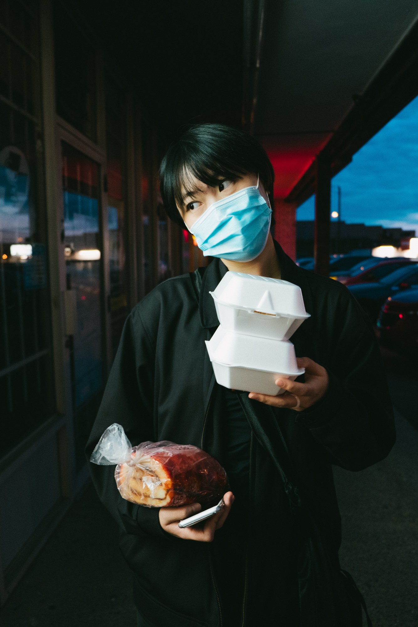 A girl wearing a mask walks along shop fronts at night carrying takeaway containers, a guava roll and her phone