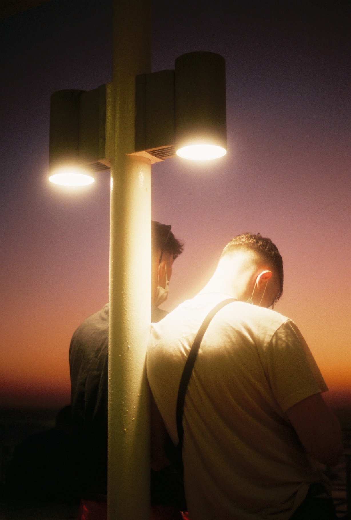 Two men watch the sunset standing against a street lamp