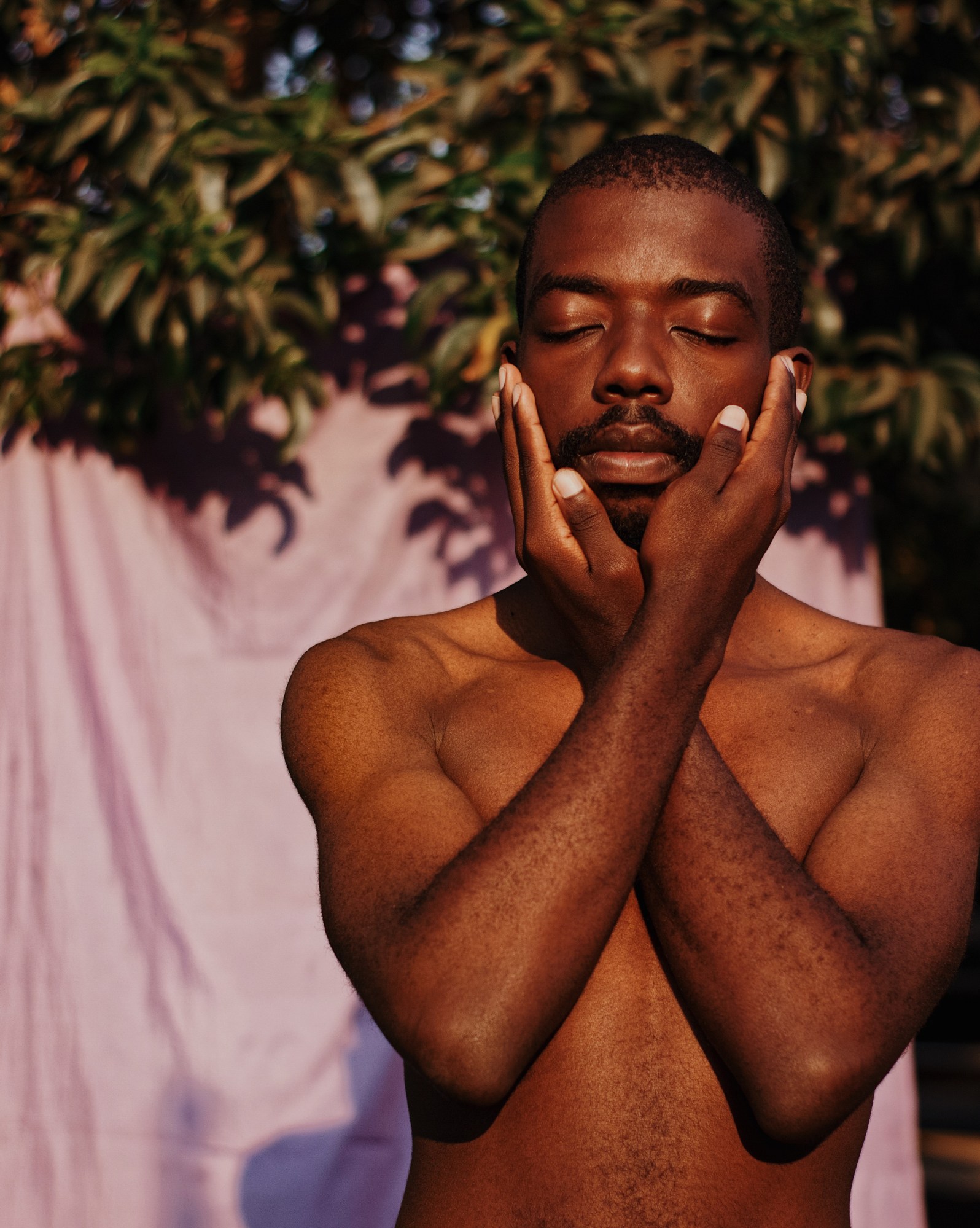 person stands with their arms crossed and hands on their face in front of bed sheets.