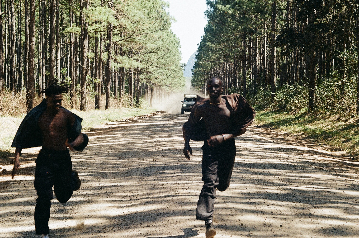 Two boys running in front of a car along a road lined with trees.