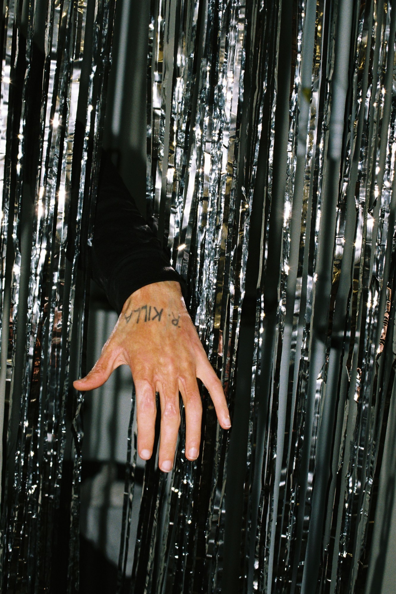 a man's hand with burn marks and an 'R. KILLA' tattoo appearing from silver streamers