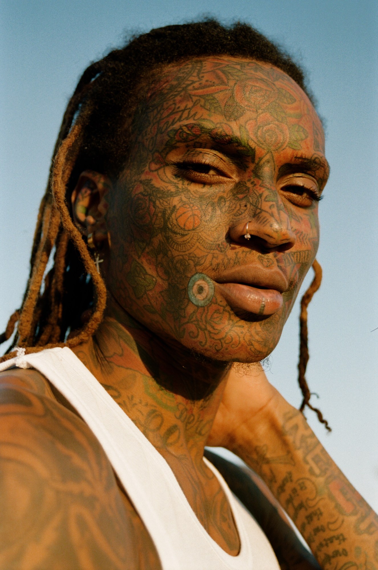 A man with tattoos covering his face and neck in a white vest top