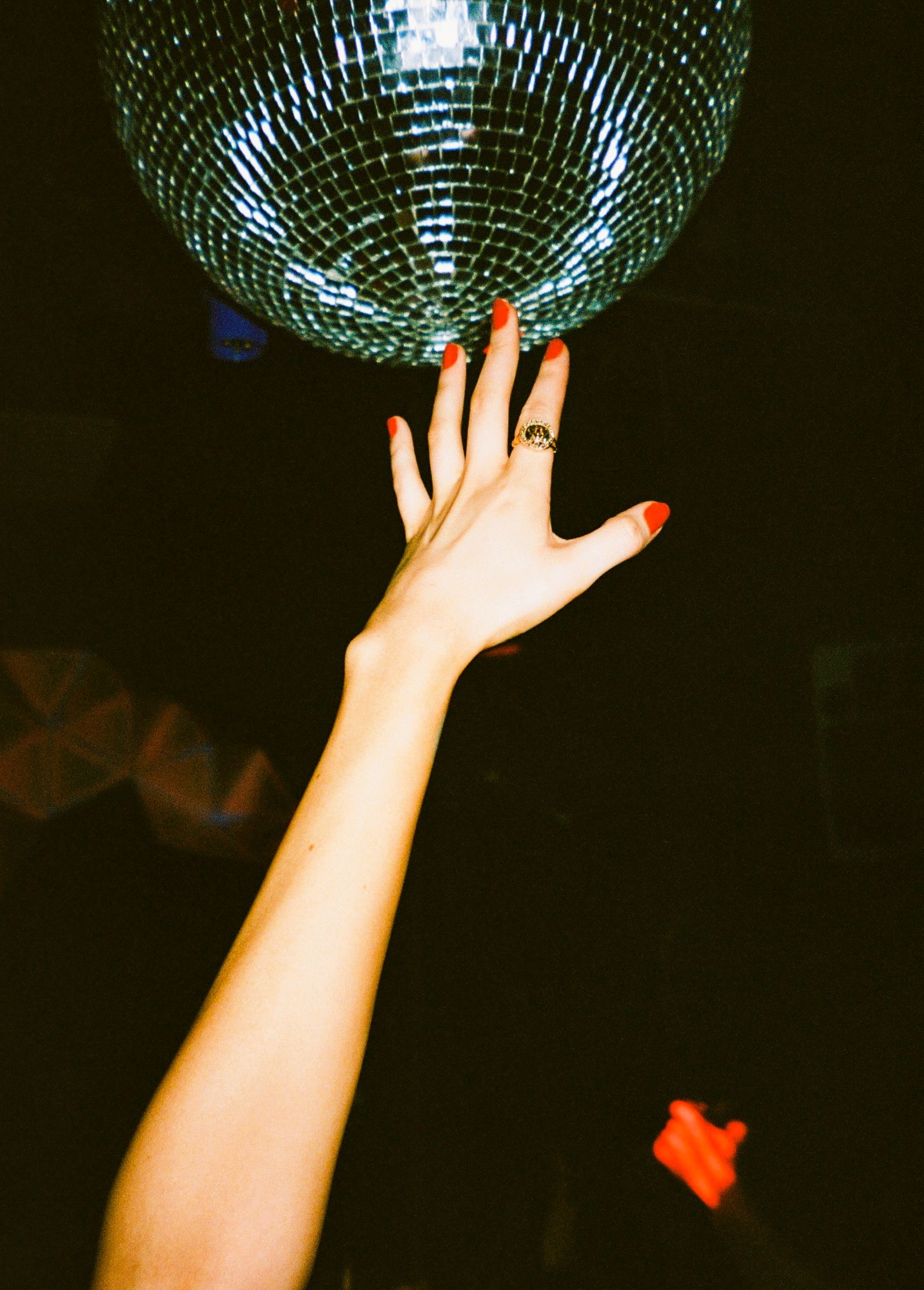 a flash photograph of a hand with red painted nails and a gold ring reaching up to a disco ball