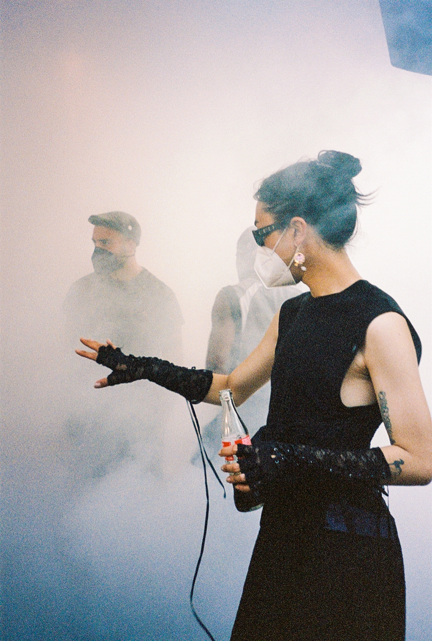 an elegant female figure in a black dress and gloves wears a mask and holds a bottle of coke as the fog of a smoke machine engulfs her