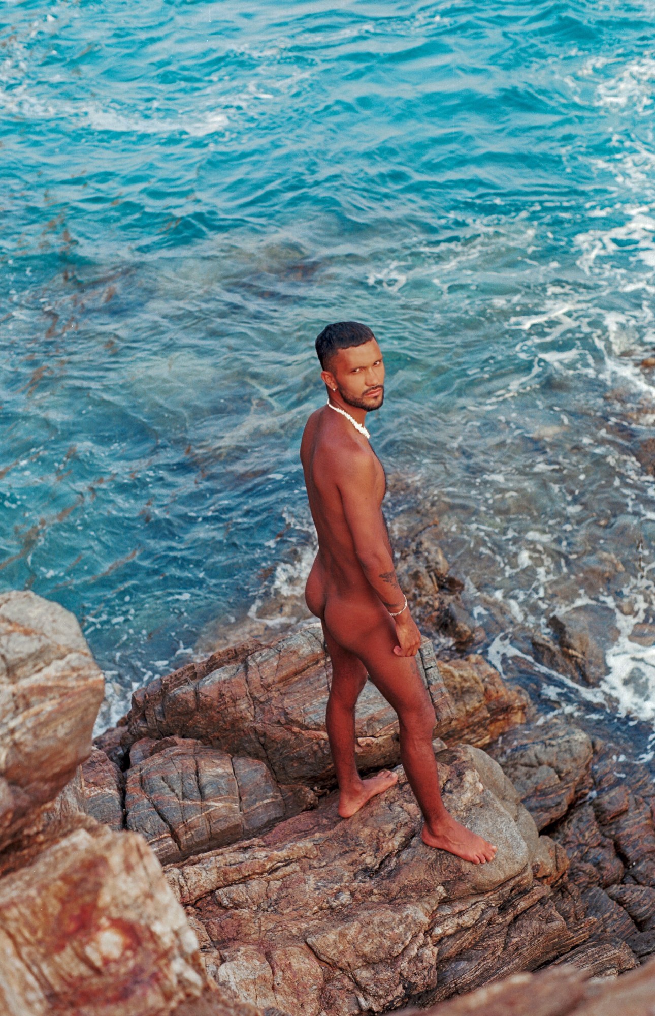 a man stands naked on rocks at the beach