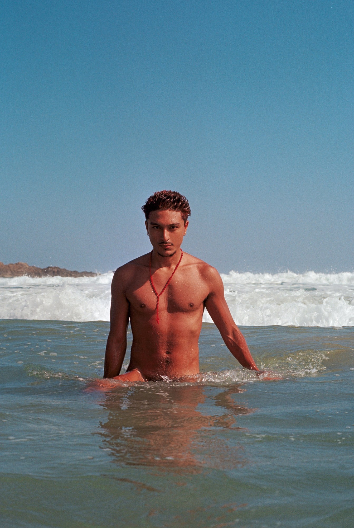 a man comes out of the water at a beach naked, wearing a crucifix