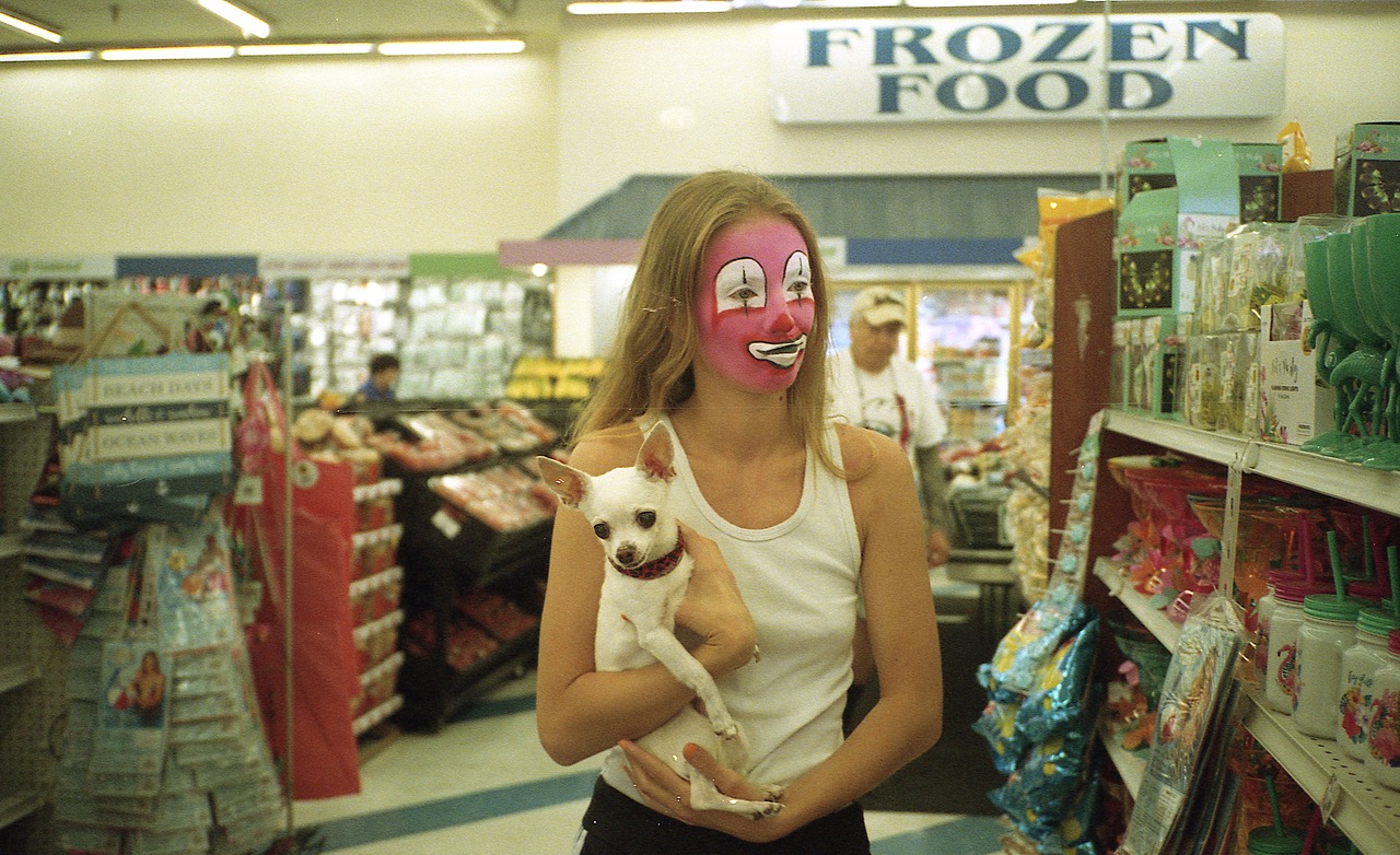 a woman wearing clown makeup holds a small dog in a supermarket