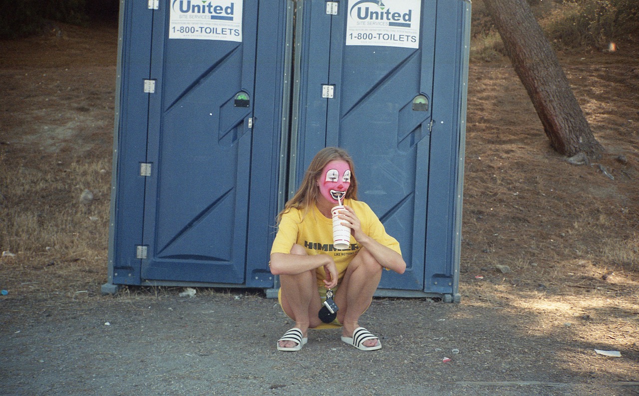 a woman in clown makeup squats and drinks from a straw in front of two portable toilets