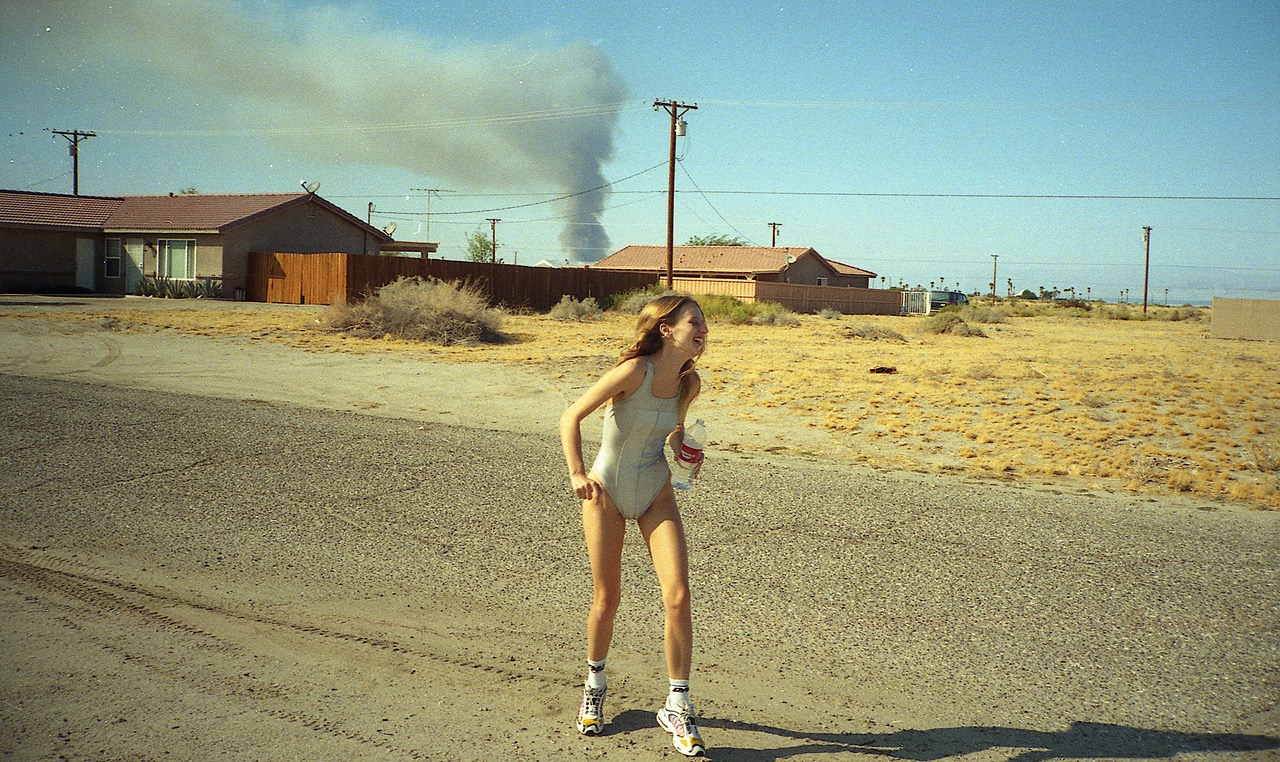 a woman in a bathing suit stands on an empty road, smoke billows in the background