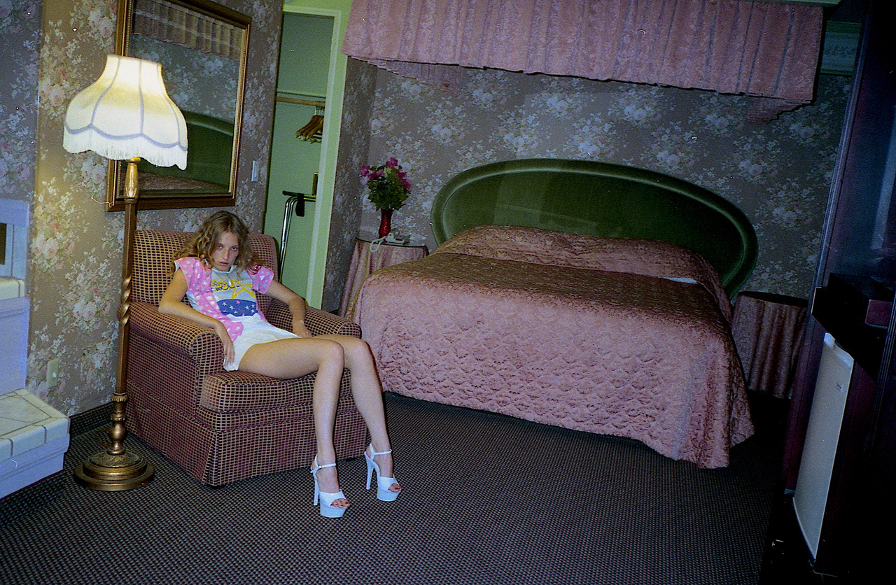 a woman wearing heels sits on an armchair in a dated hotel room