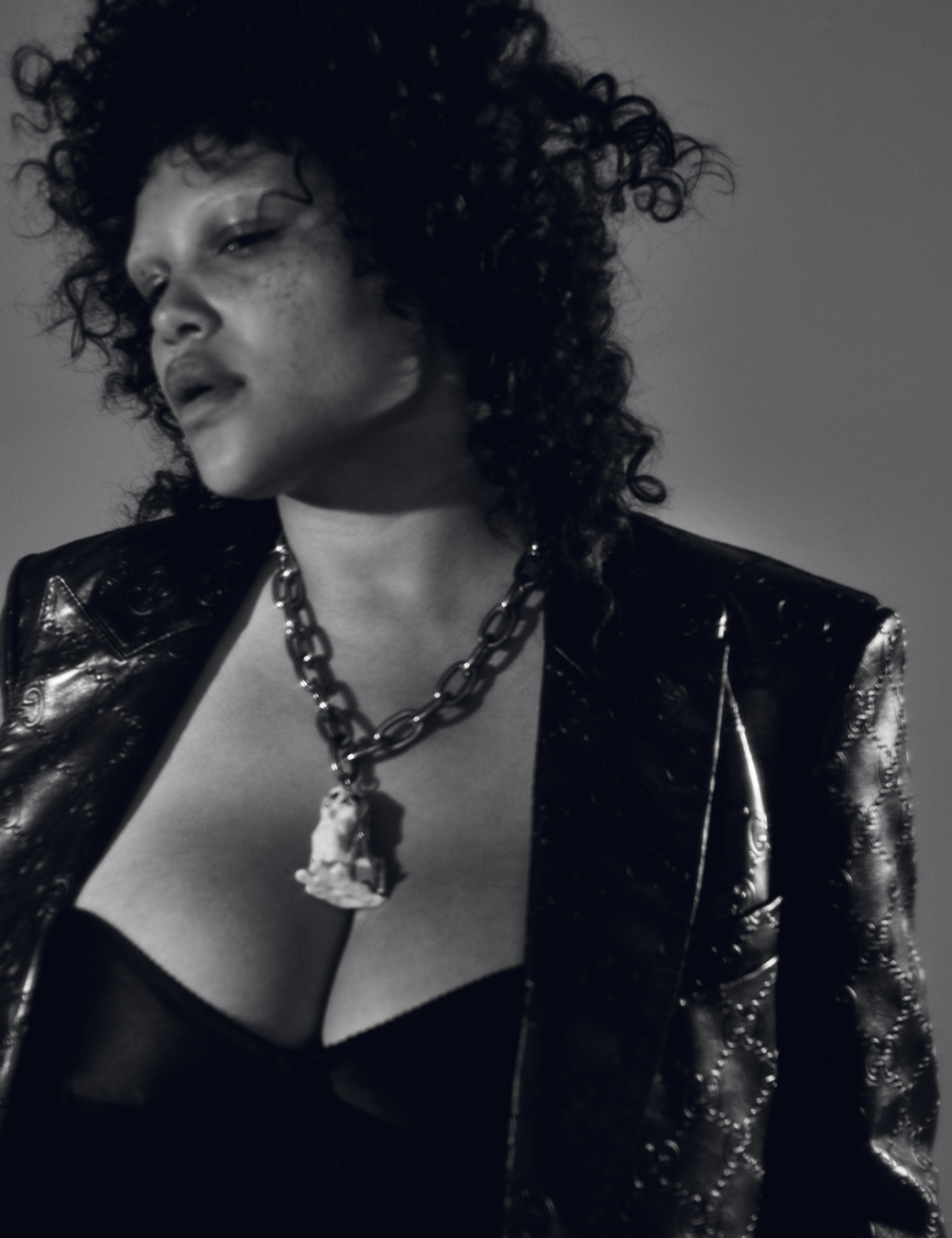 model is shot from the chest up in a corset and leather blazer, her hair is styled curly and a large chain necklace hangs from her neck