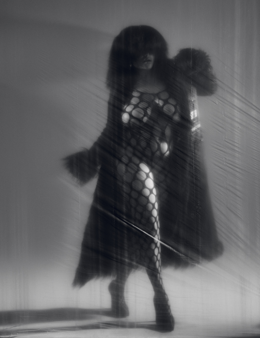 model walks behind a thin plastic sheet in a patterned bodysuit, her eyes obscured by a heavy fringe