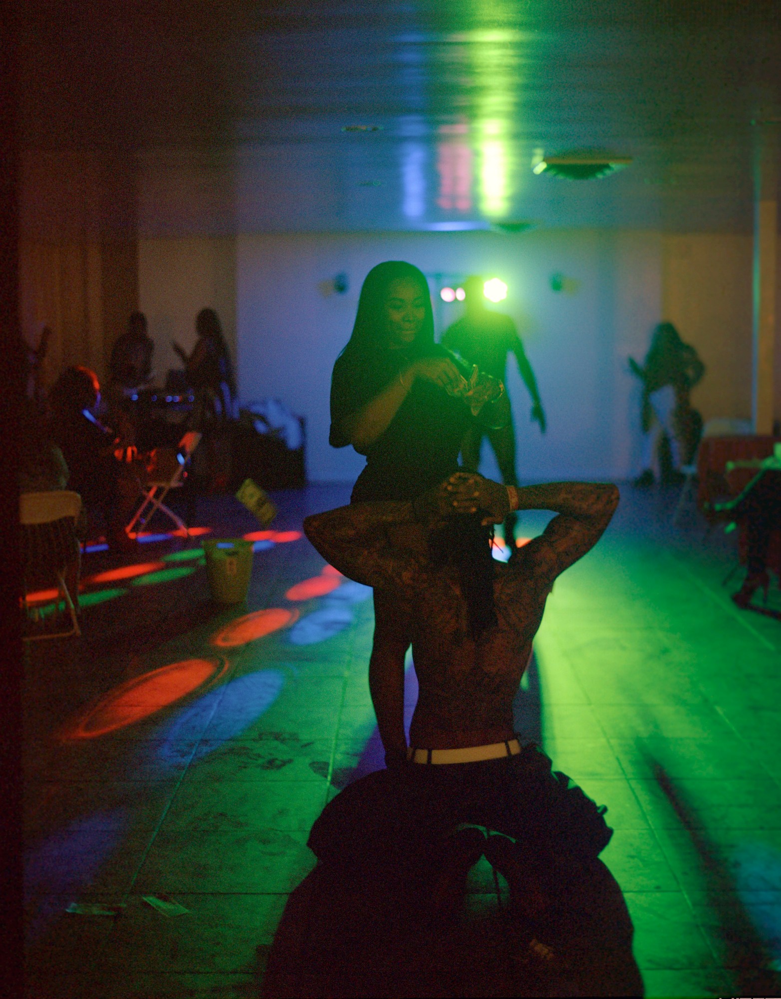 a tattooed man kneels before a woman on a neon-lit dance floor. she films him on her phone