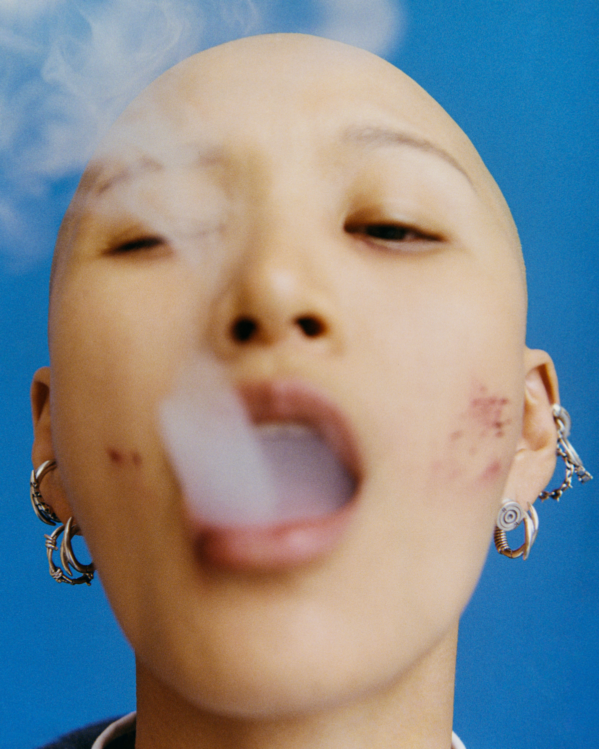 a model with lots of silver hooped earrings and a shaved head blows smoke from their mouth, obscuring part of their face