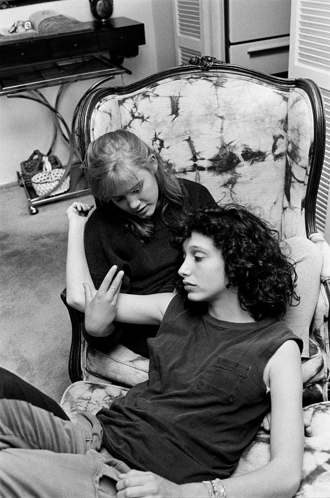 a young woman in a tank top leans against another woman sat on a chair