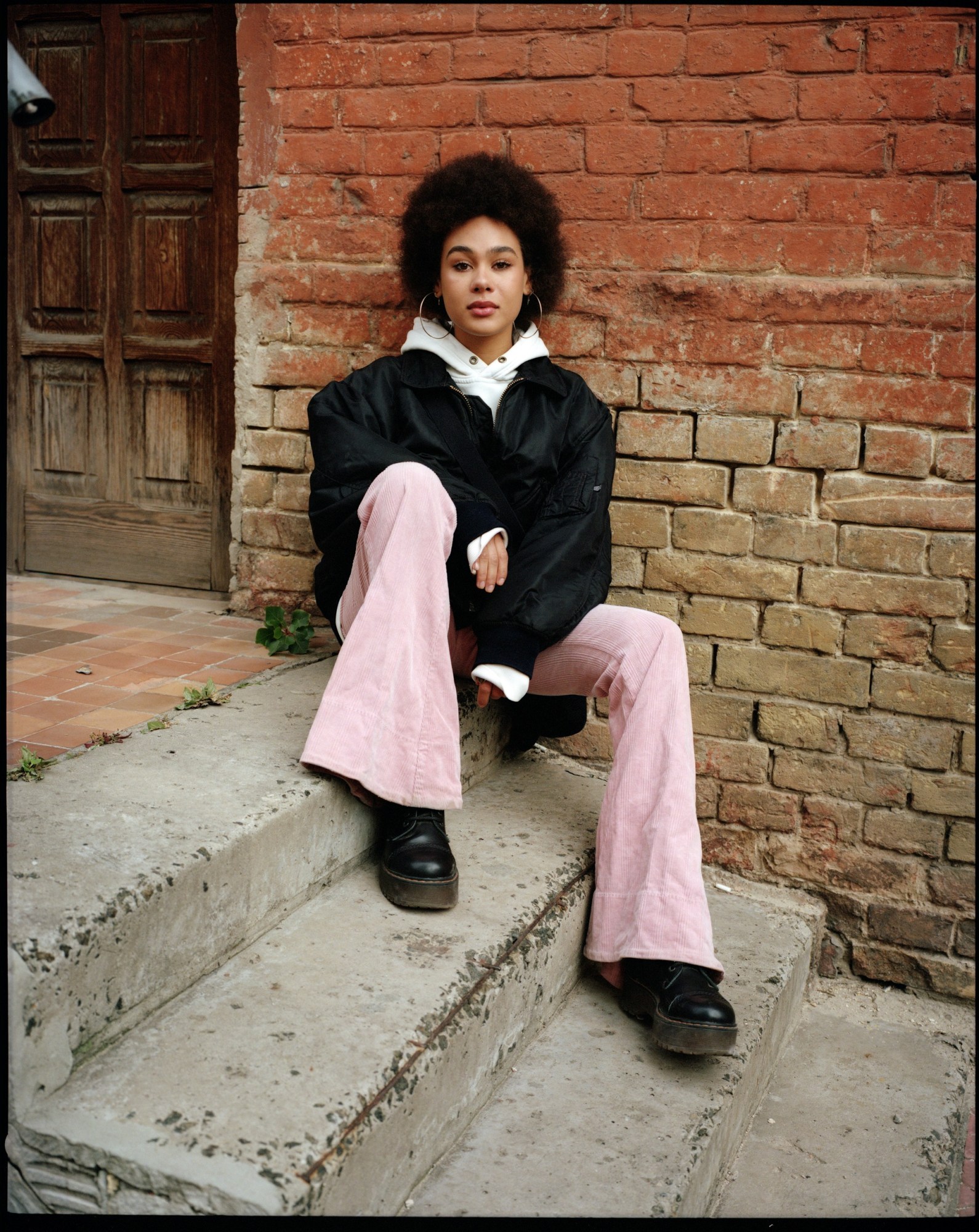 a woman with an afro, pale pink cords and a bomber jacket sits on some outside steps