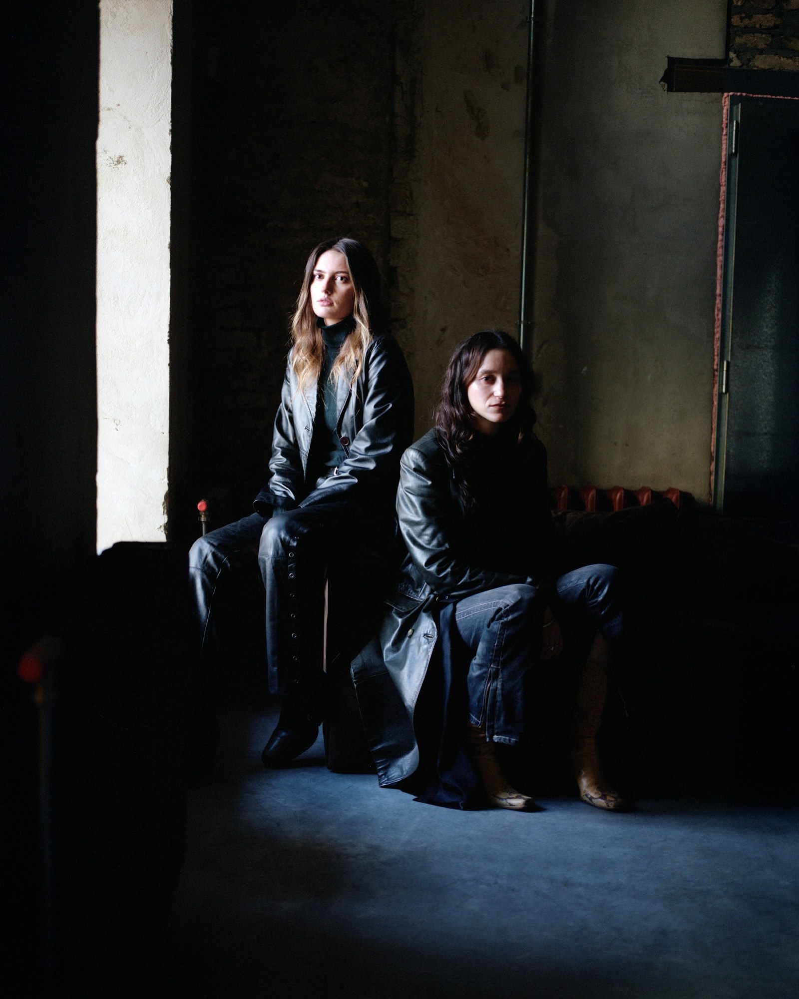 two women wearing leather jackets sit in a dark room by a window, light shines on their faces