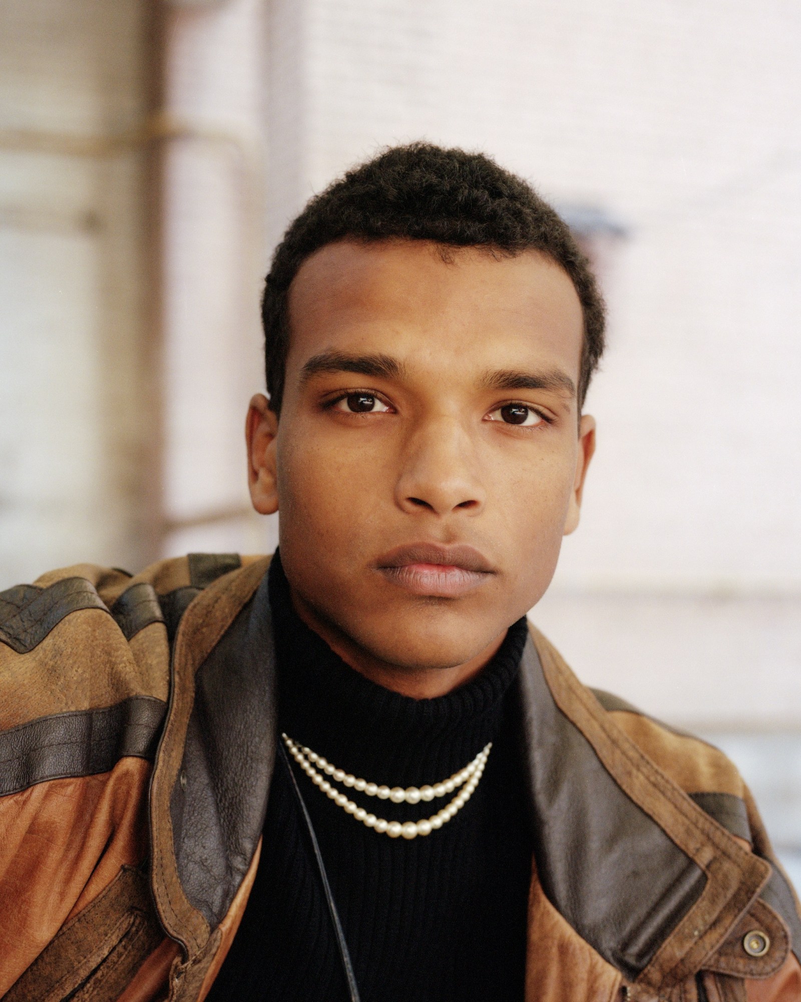 a young man wearing a brown suede jacket and pearls looks into the camera