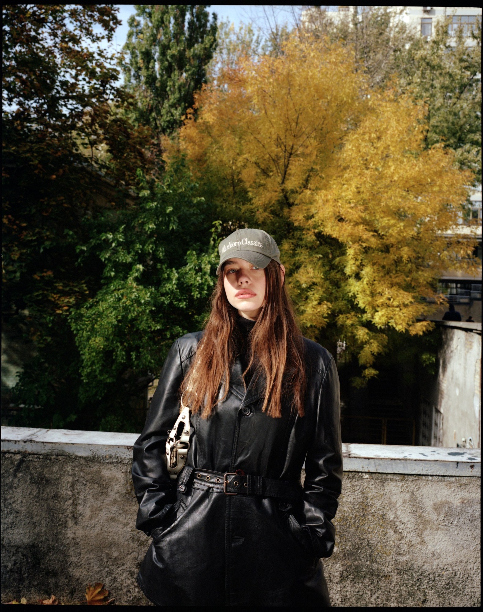 a woman wearing a belted leather jacket and a grey cap stands against a concrete wall. green and yellow trees are behind her