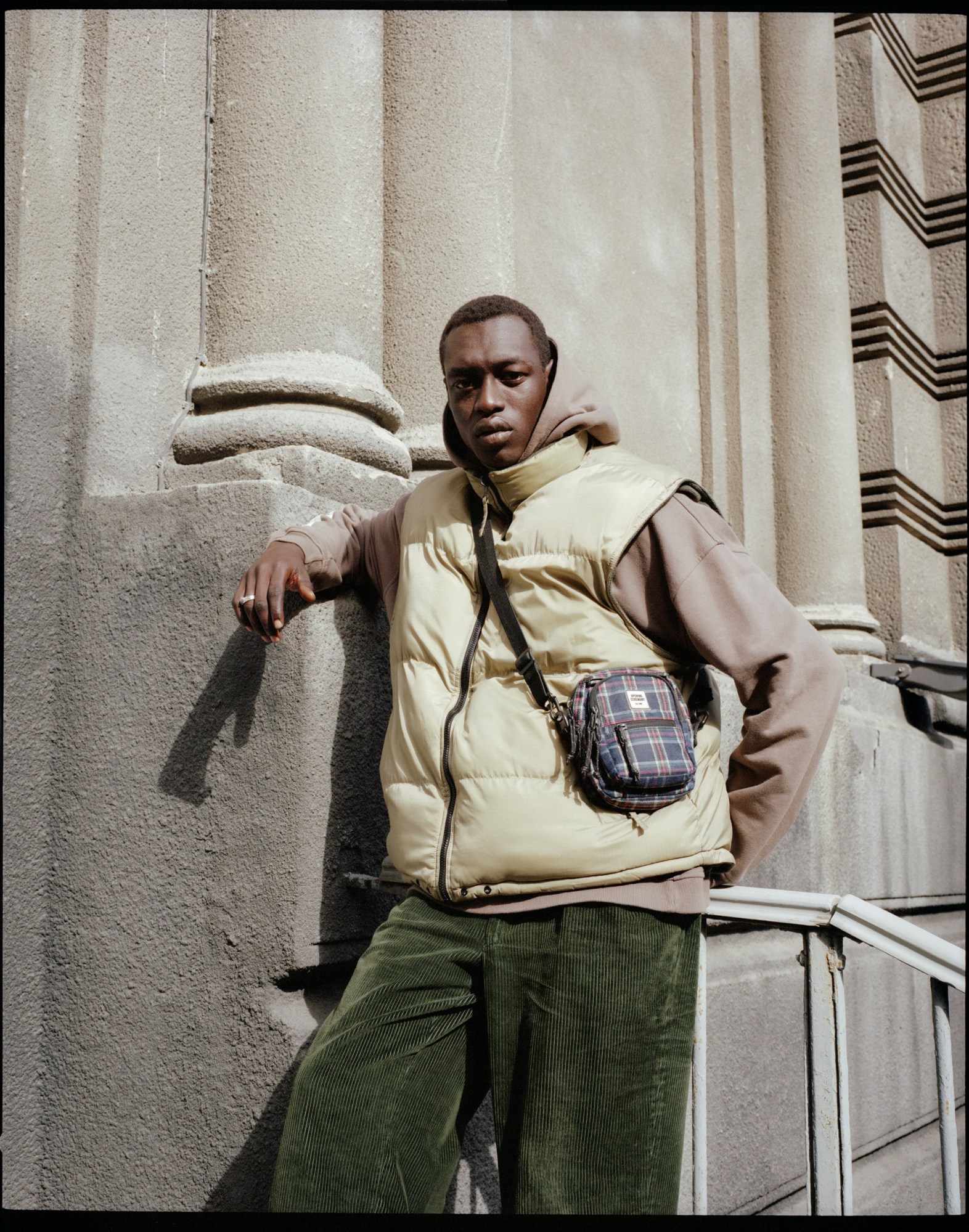 a young male model wears green cords and a gilet over a hoodie. he leans against a concrete building and looks to camera