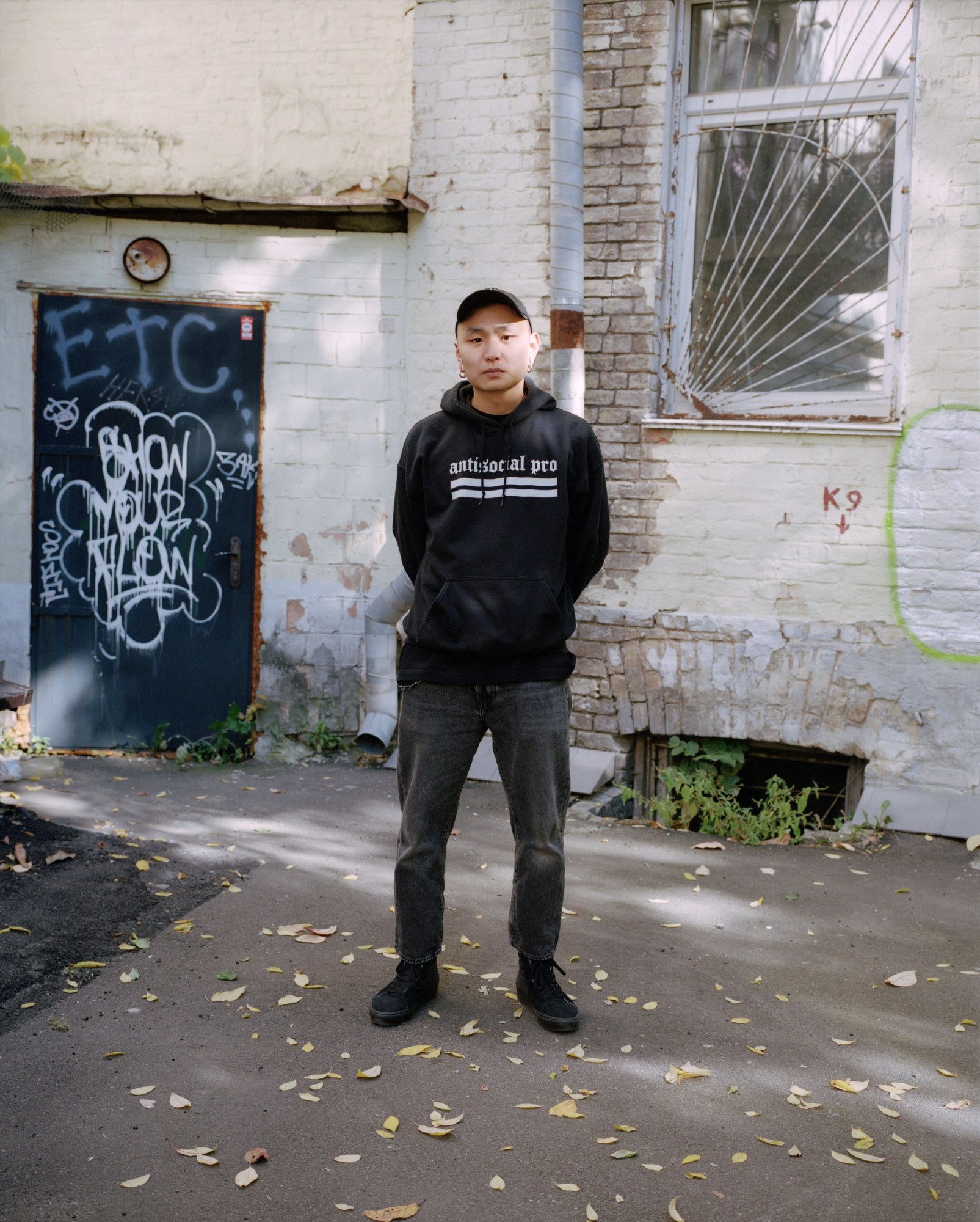 a man wearing a hoodie with the words 'antisocial pro' stands in a scruffy courtyard with his hands behind his back, looking to camera