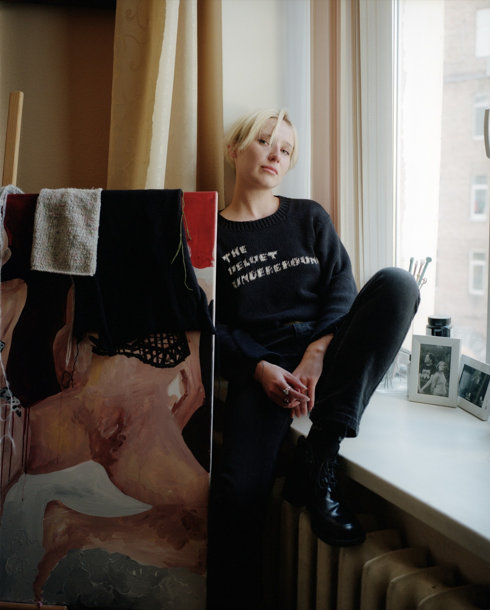 a woman with short blonde hair sits on a windowsil next to a painting of a person sitting nude with their legs open