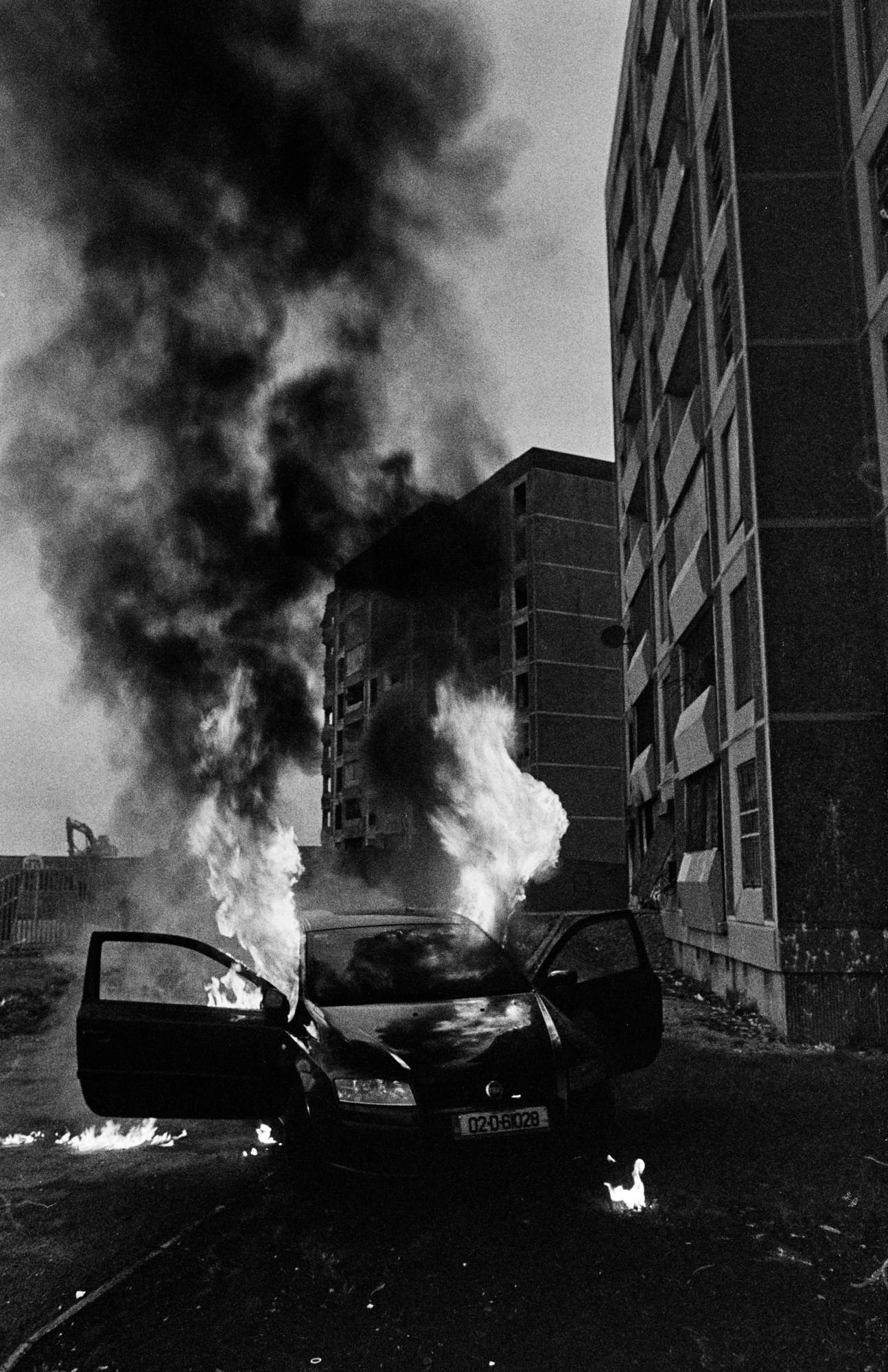 a car on fire in front of a dilapidated tower block