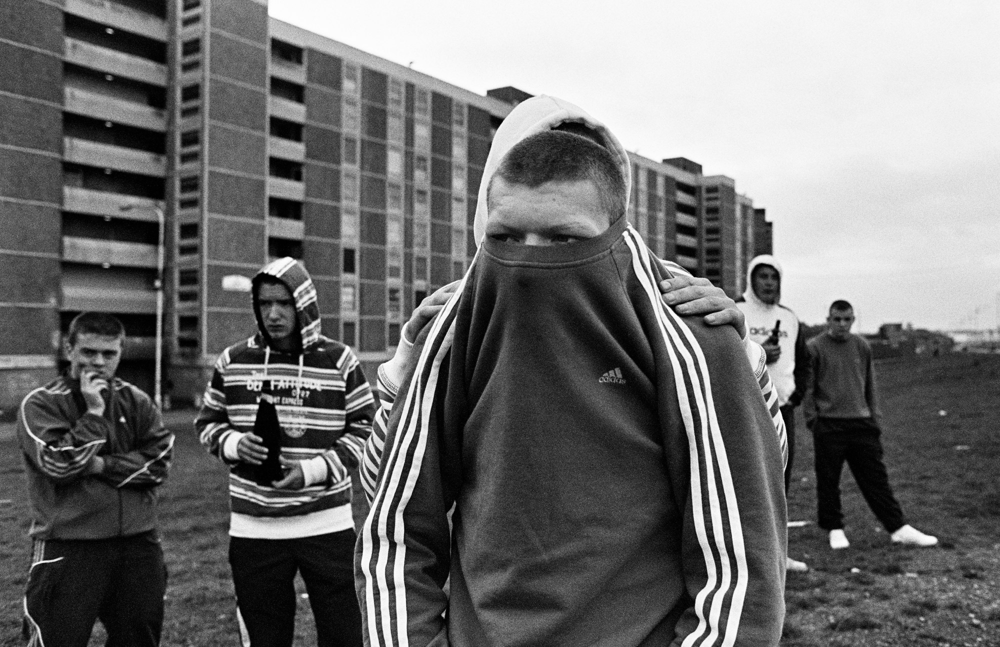 a young man has a jumper lifted up to his eyes, men in tracksuits stand behind him