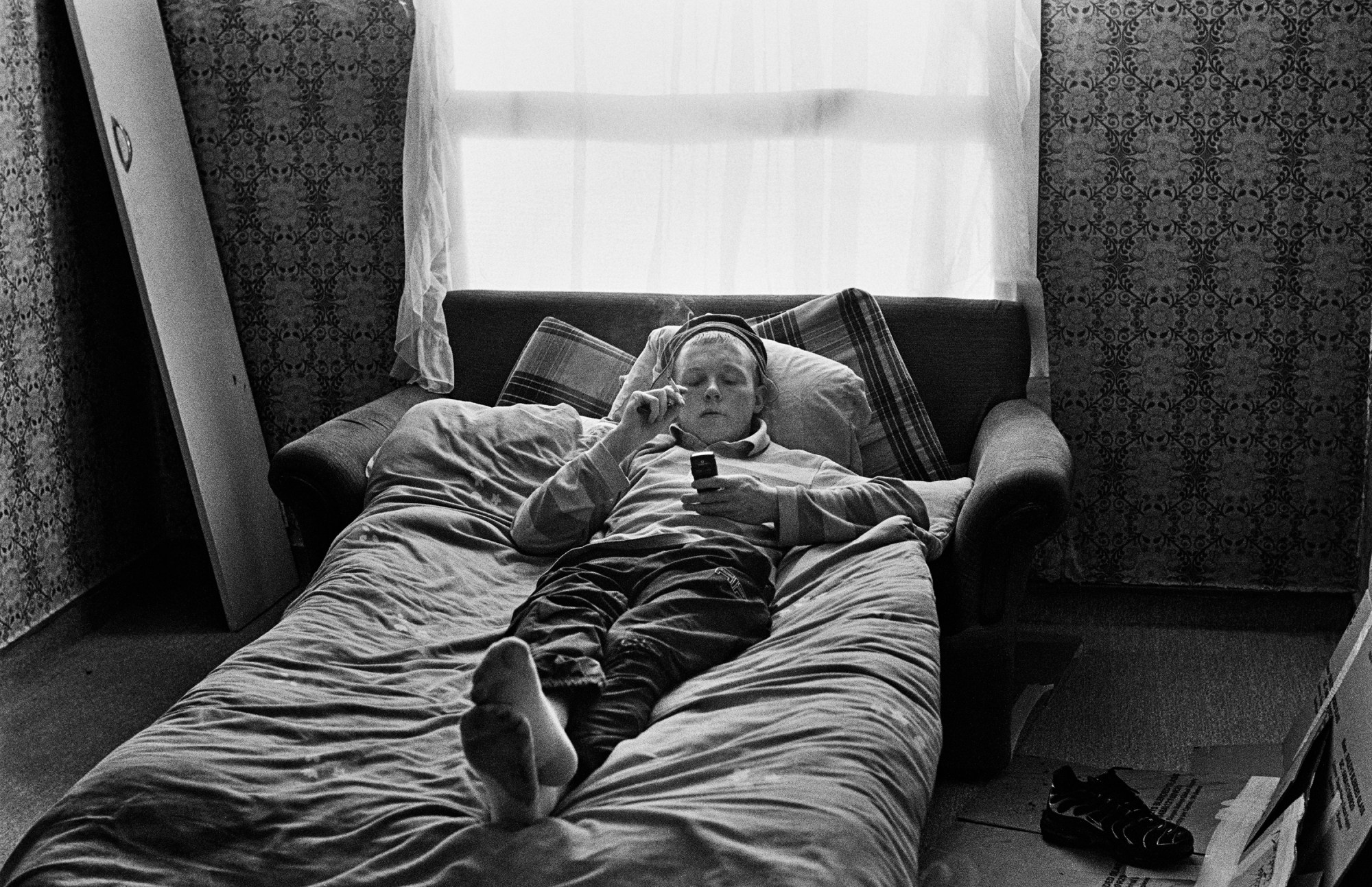 a young man lies on a sofa bed, smoking and looking at a mobile phone