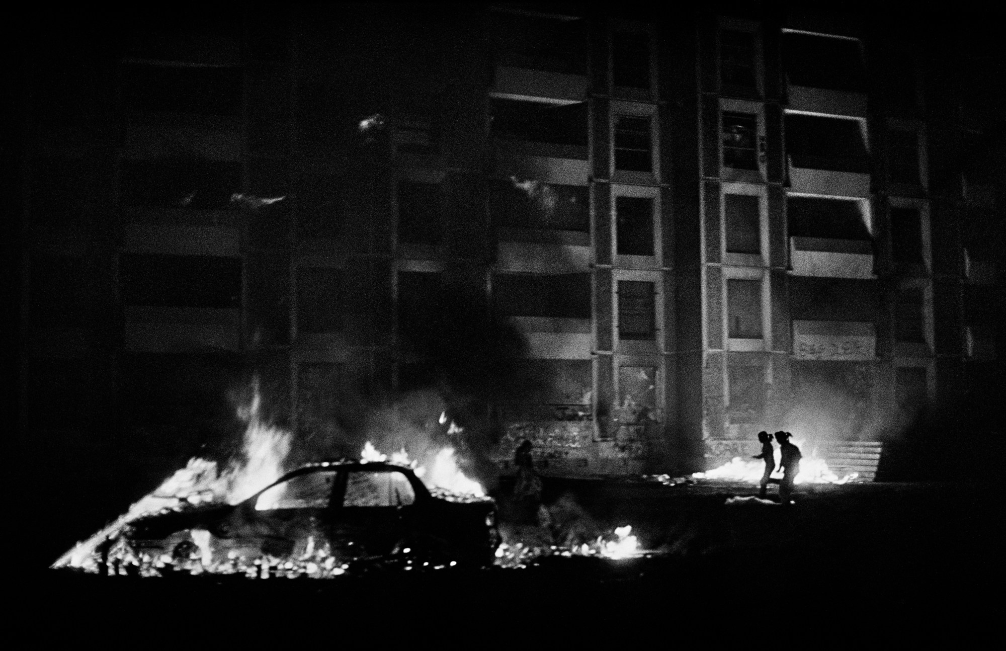 a car on fire and two silhouettes running against the flames