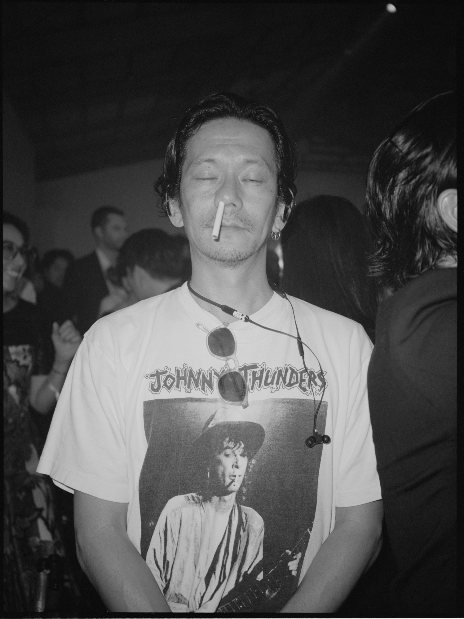 a man wearing a johnny thunders band tshirt stands at a gig looking to camera with a cigarette up one nostril