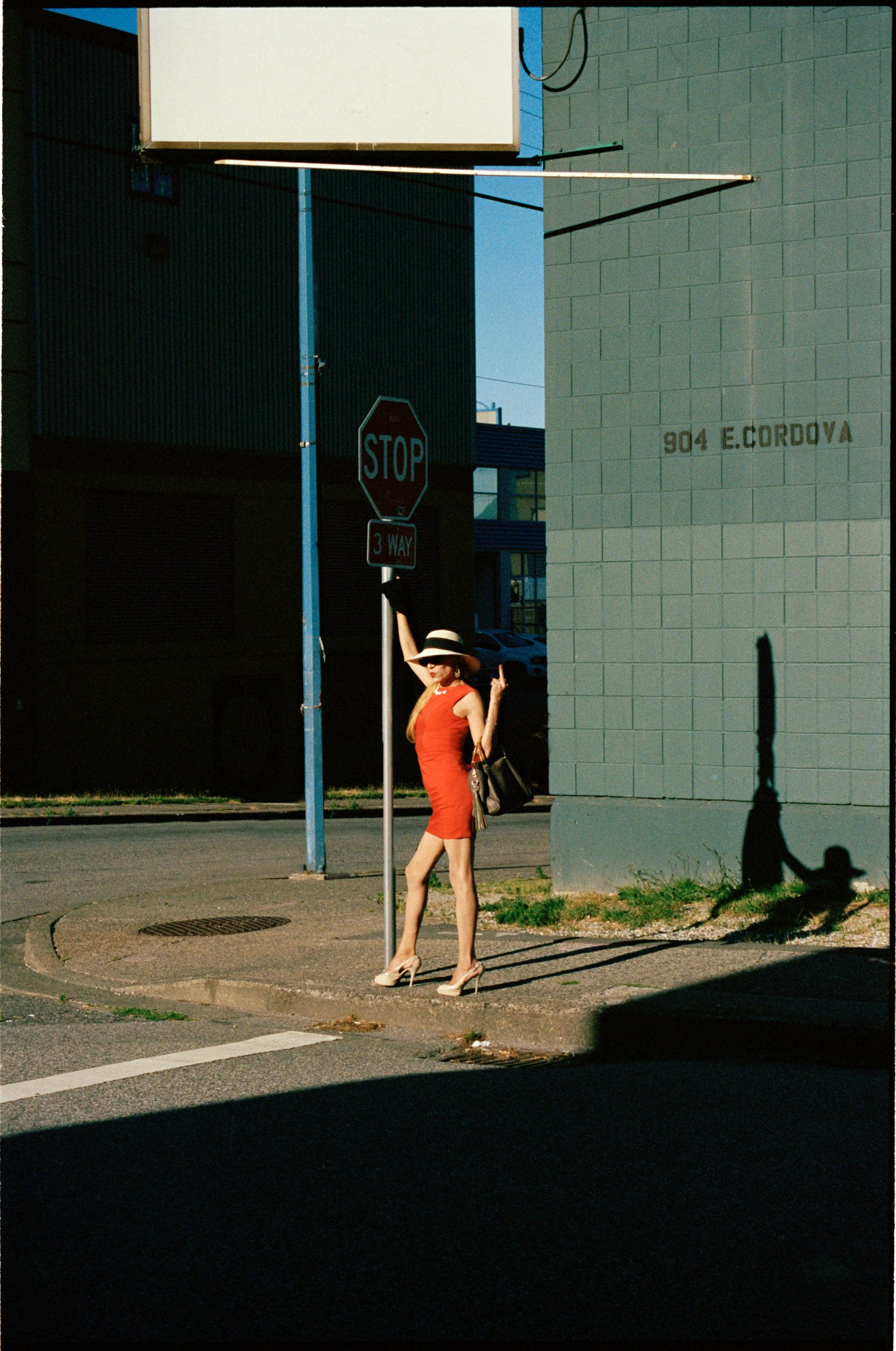 a woman in a red dress and a trilby hat stands waiting to cross the street with their middle finger up