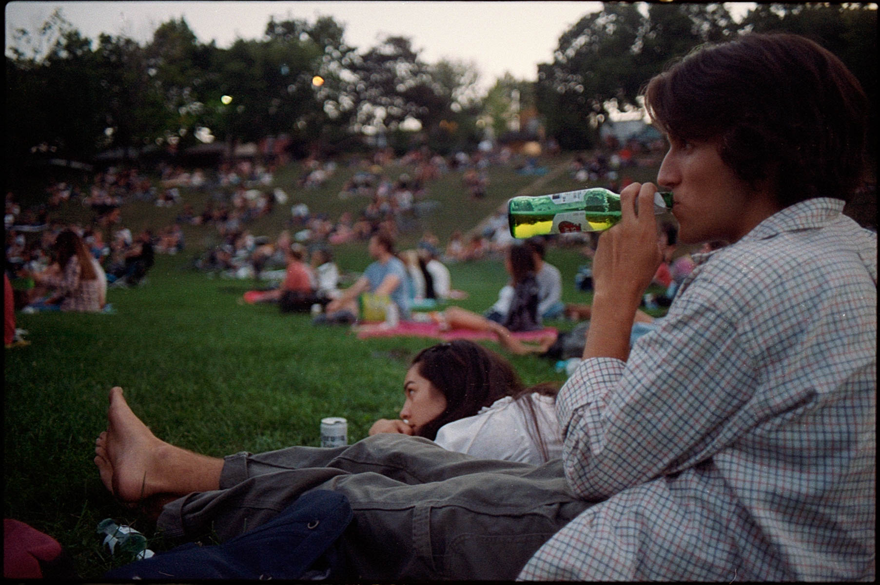 a young man in a park drinks beer, surrounded by people