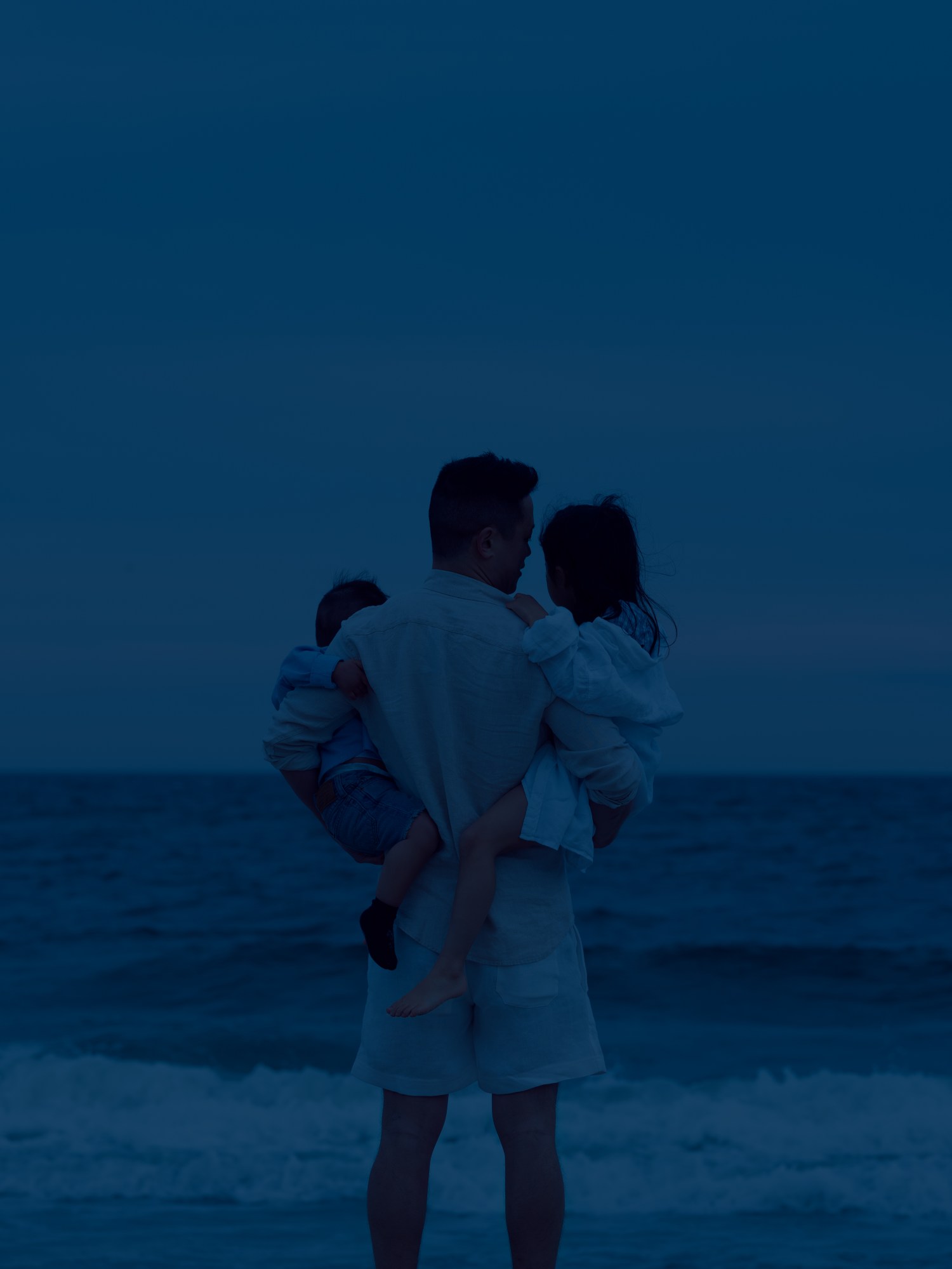 A father holds his two daughters at night under the moonlight by the incoming ocean tides.