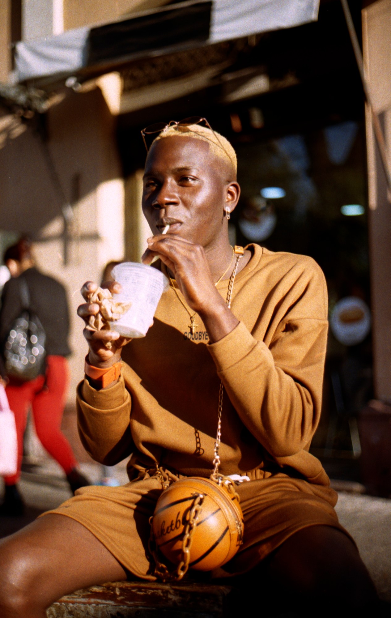 Person in brown sweats and a basketball bag drinks a milkshake while sitting on the side of the road.