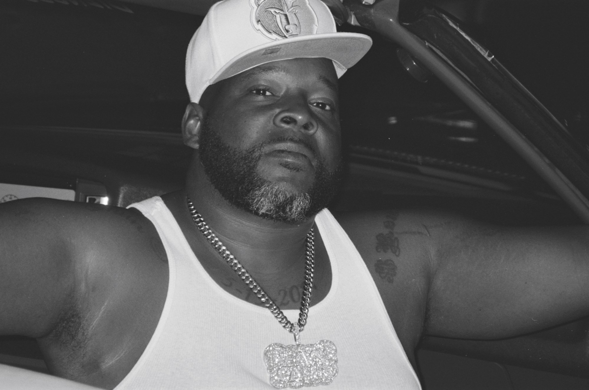 A portrait of a man sitting in a car, wearing a white hat and a diamond chain.