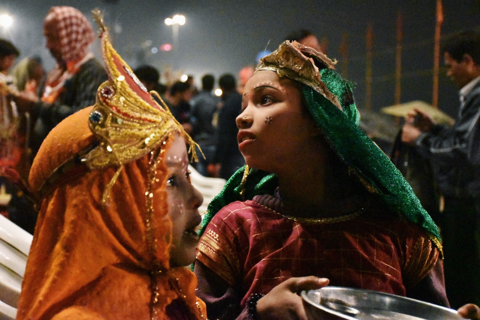 Two kids dressed up and with face paints and jewels walk through a crowd.