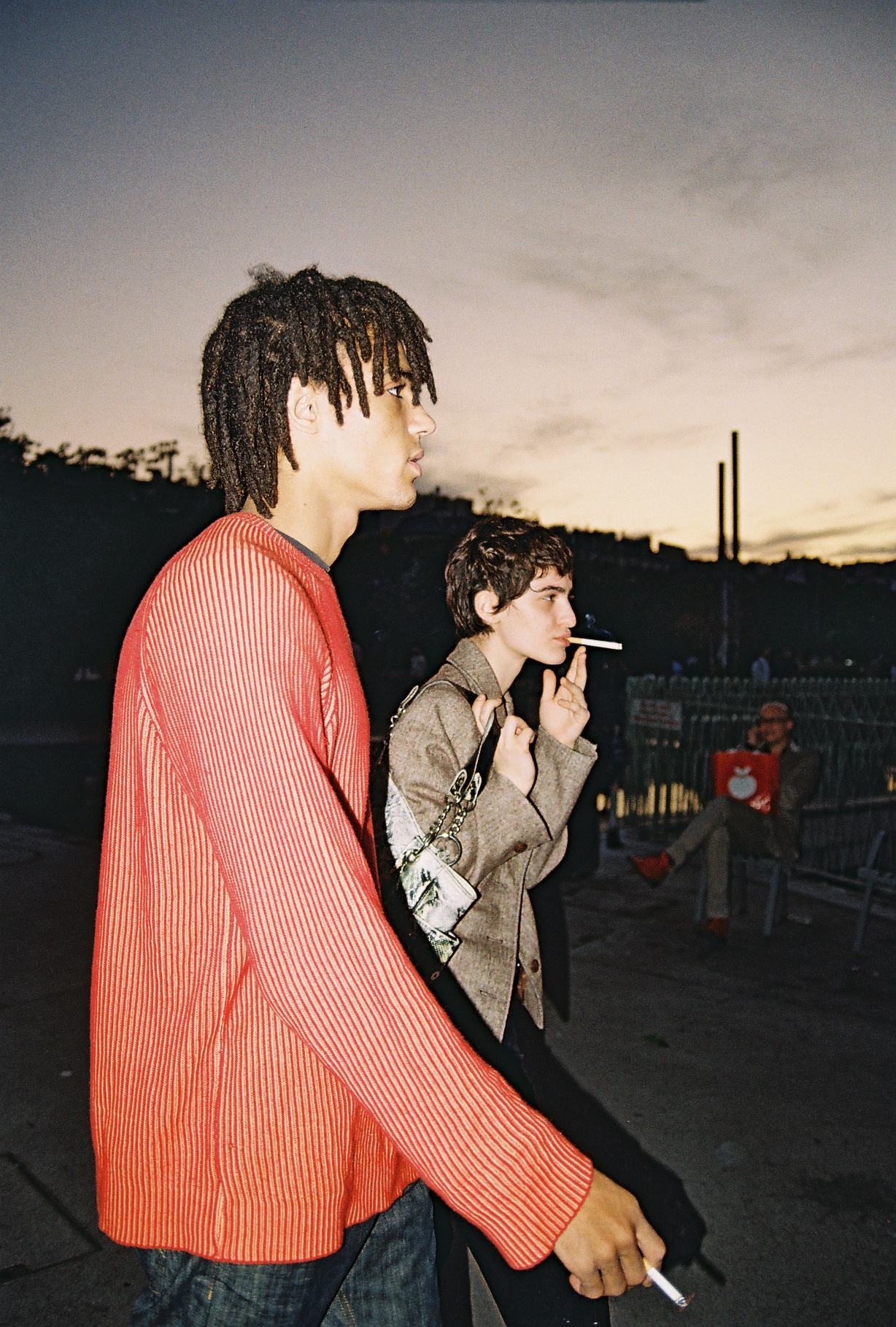 Two people walk through the street smoking.