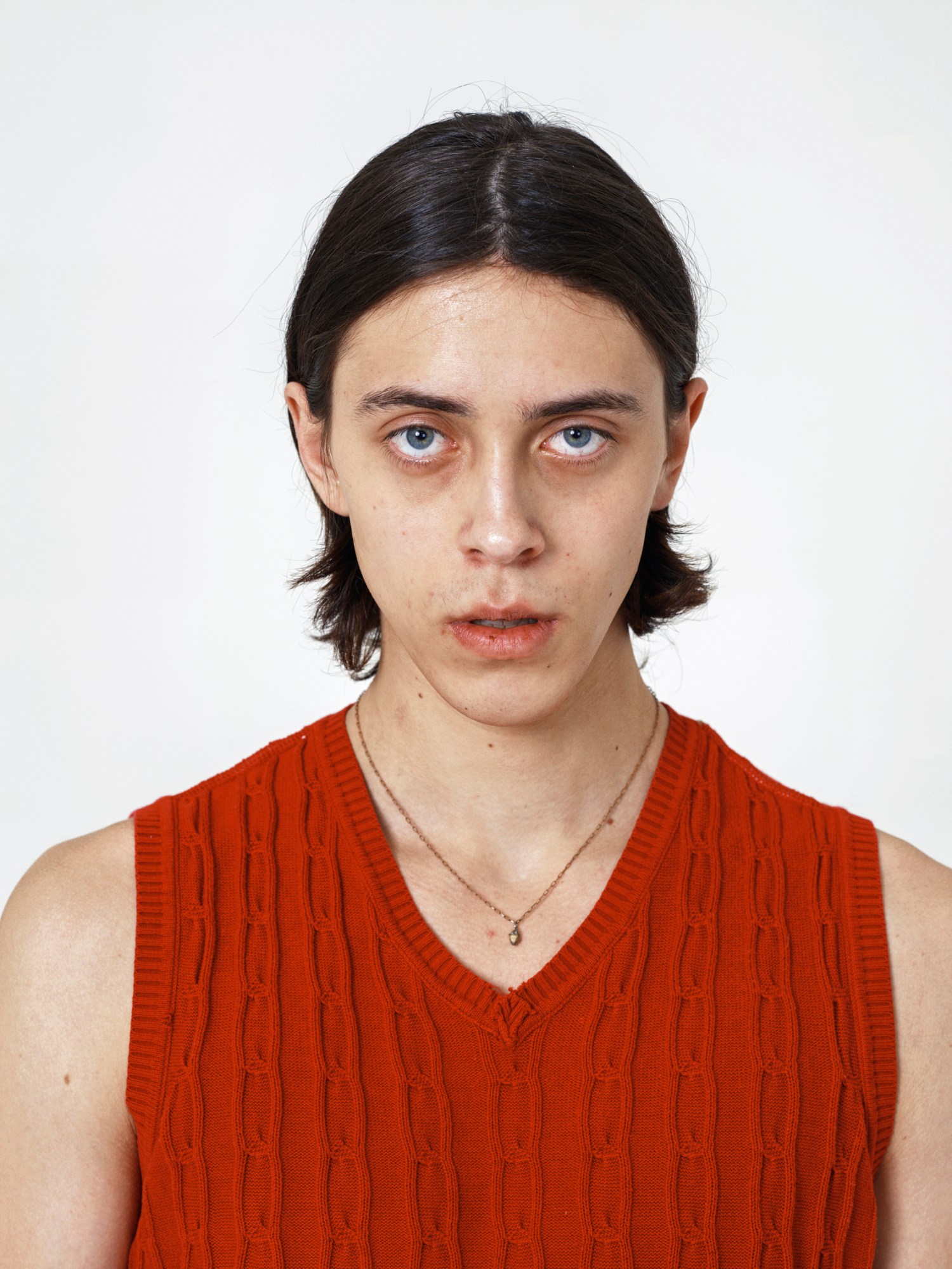 Man with black long hair pushed behind his ears wears a thin gold chain and red jumper vest.