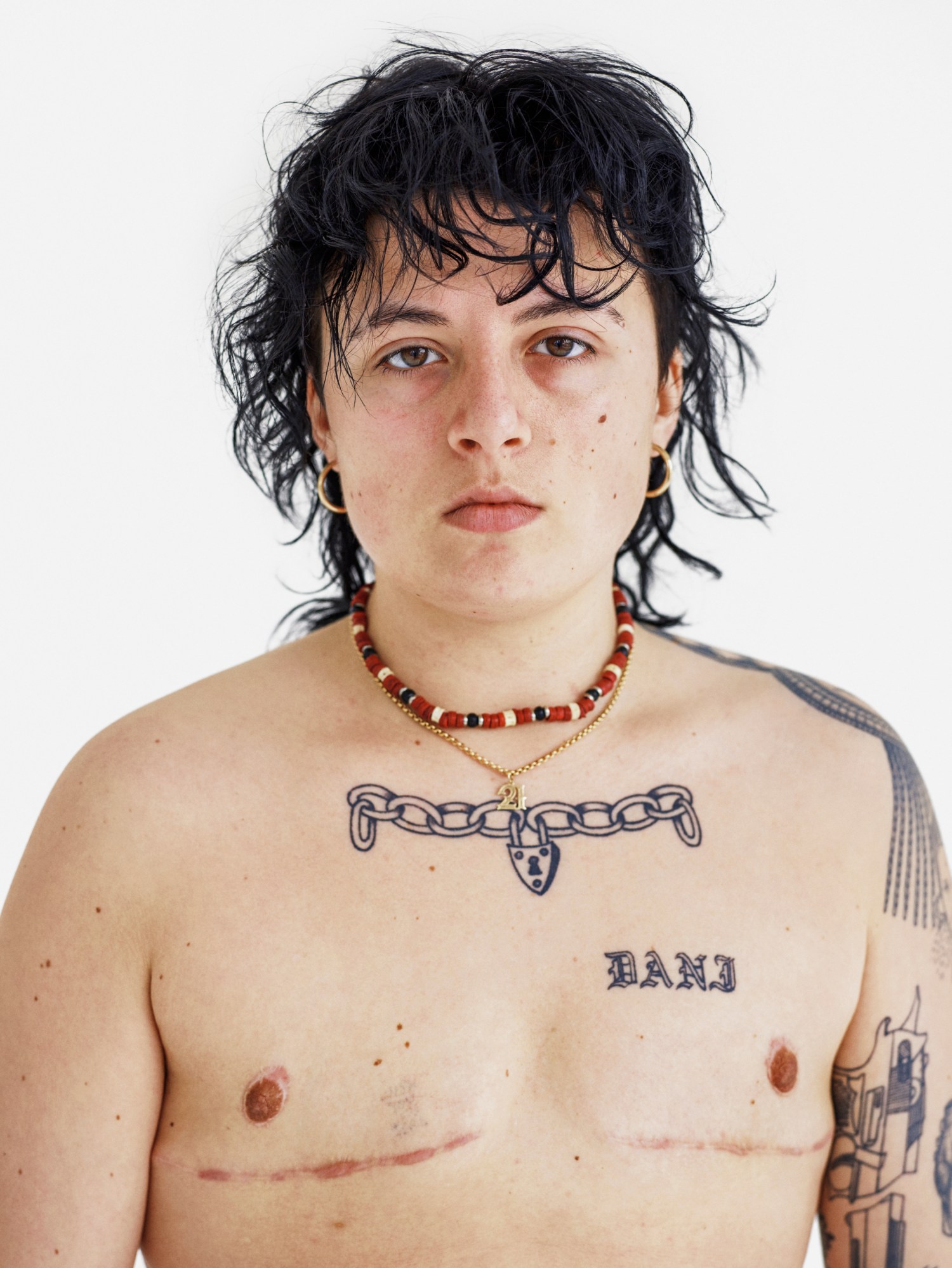 A topless man with black wavy hair, a fringe and tattoos on chest and one arm wearing a beaded necklace and a gold chain with a 21 pendant.