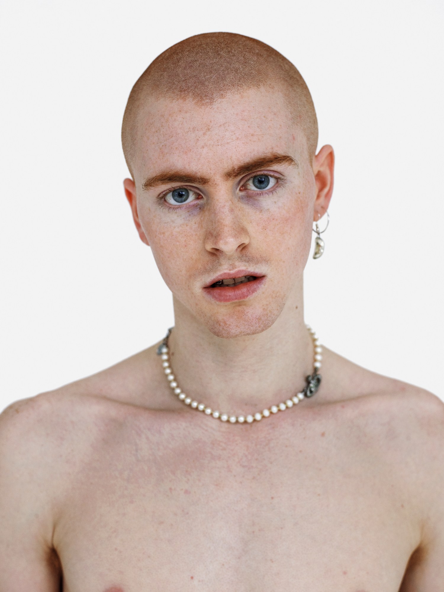 A topless man with ginger shaved hair wearing a pearl necklace and a hoop earring.