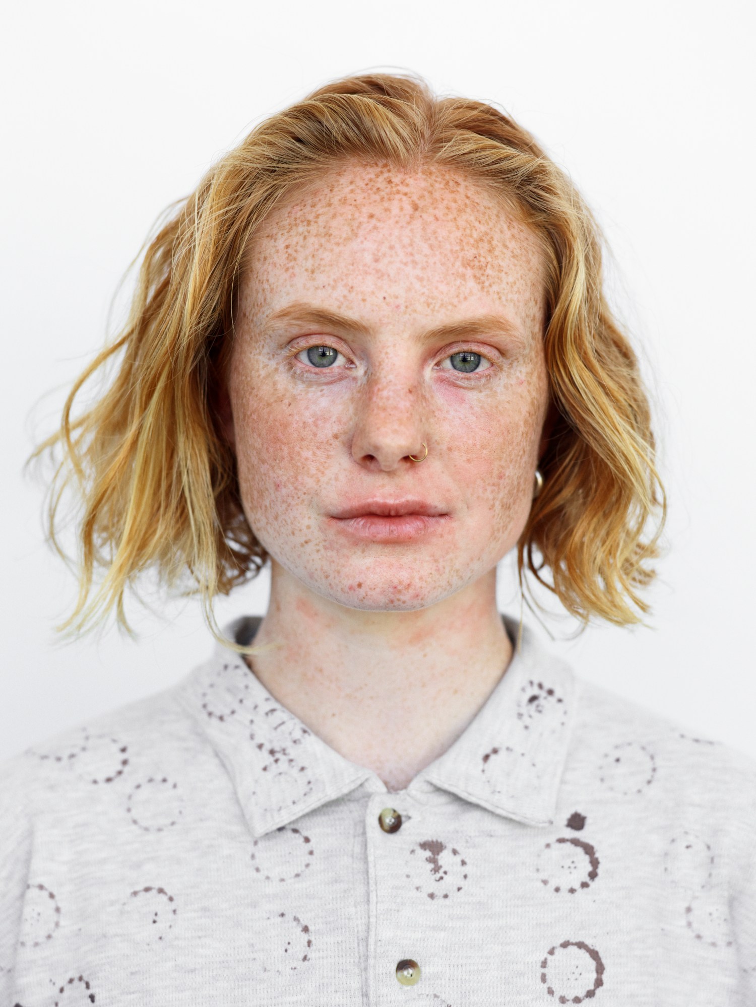 A woman with freckles, a nose piercing and a blonde bob hair cut wearing a grey printed polo top